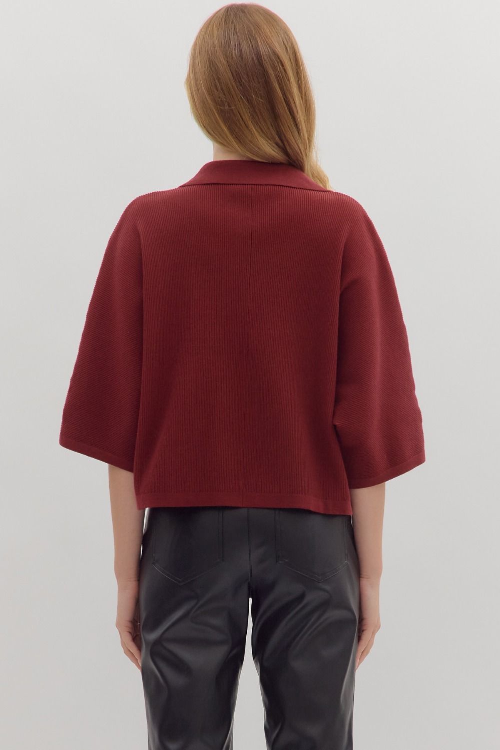 Back view of woman wearing slightly cropped ribbed 3/4 sleeve burgundy knit sweater and black pants on a white background