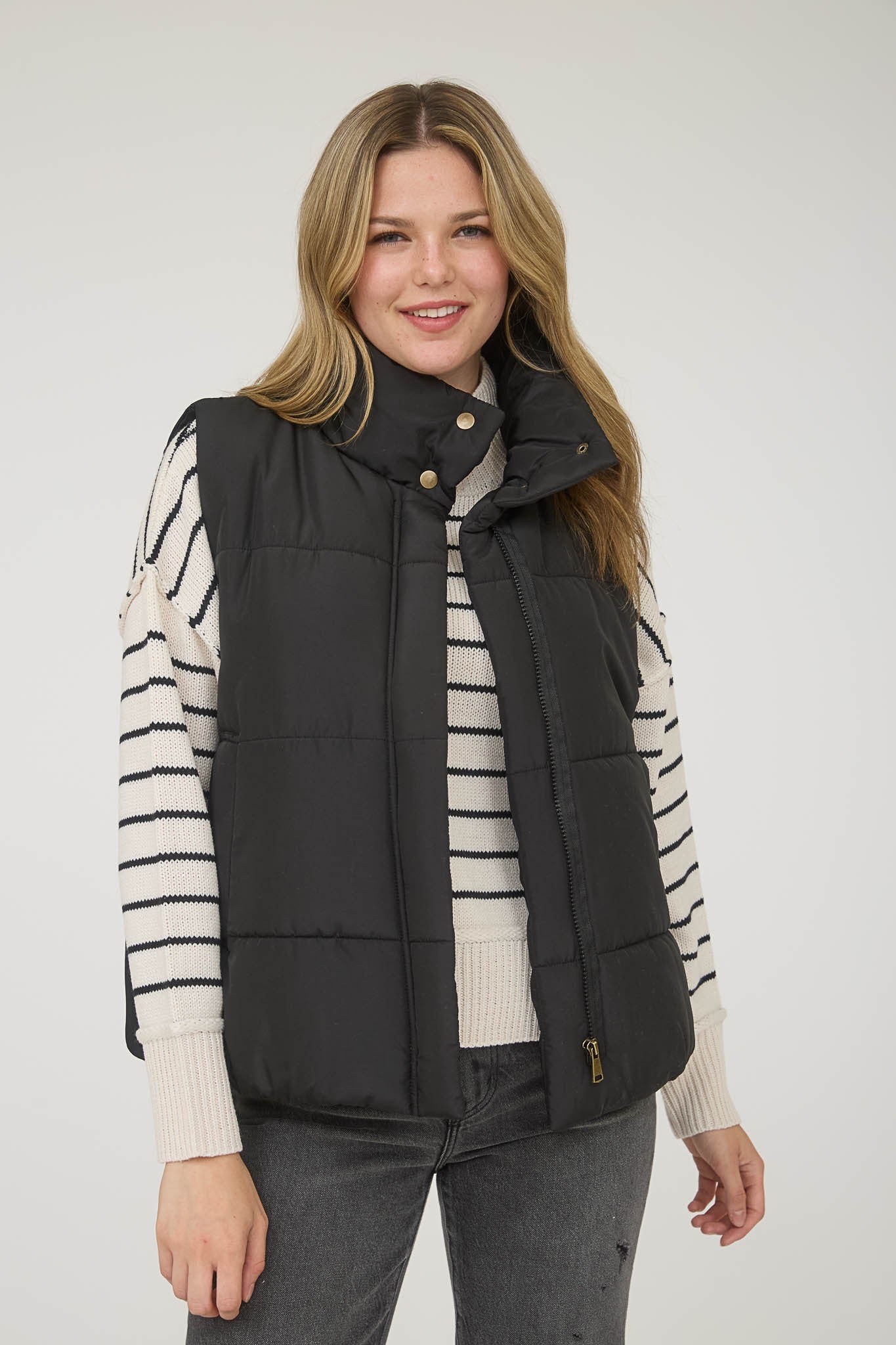 Woman wearing a black puffer vest with zipper closure over a striped sweater on a white background