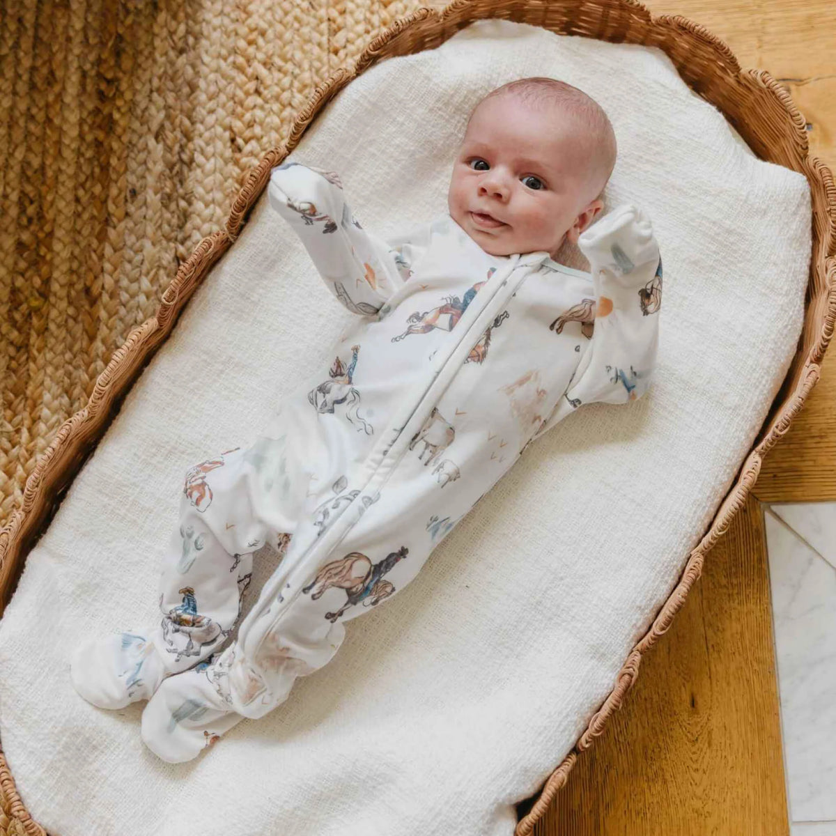 Baby in a horse and rider patterned onesie lying on a white blanket in a wooden crib.
