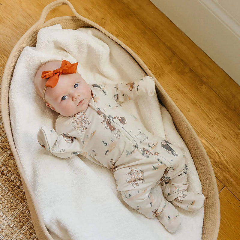 Baby in a Moses basket with a horse pattern footie outfit and orange bow on a wooden floor.