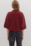 Back view of woman wearing slightly cropped ribbed 3/4 sleeve burgundy knit sweater and black pants on a white background