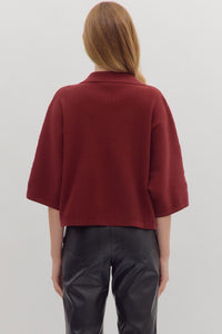 Back view of woman wearing slightly cropped ribbed 3/4 sleeve burgundy knit sweater and black pants on a white background