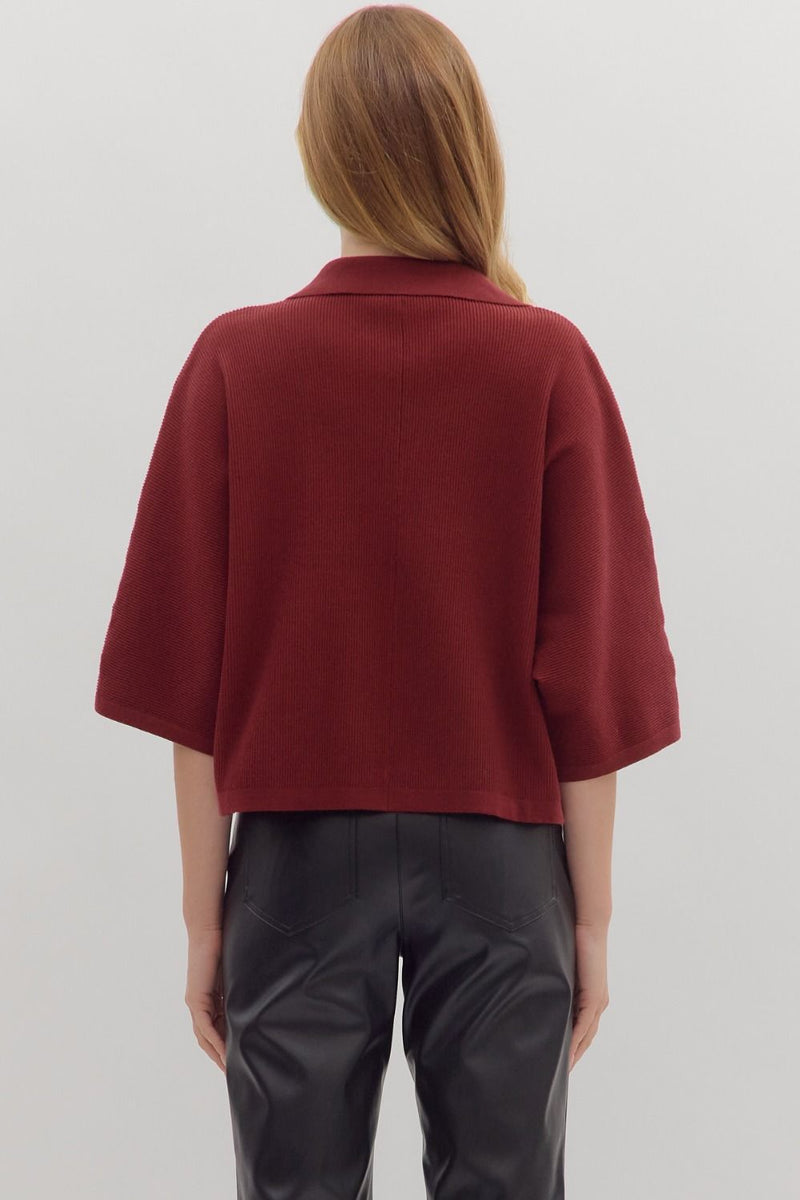 Back view of woman wearing slightly cropped ribbed 3/4 sleeve burgundy knit sweater and black pants on a white background